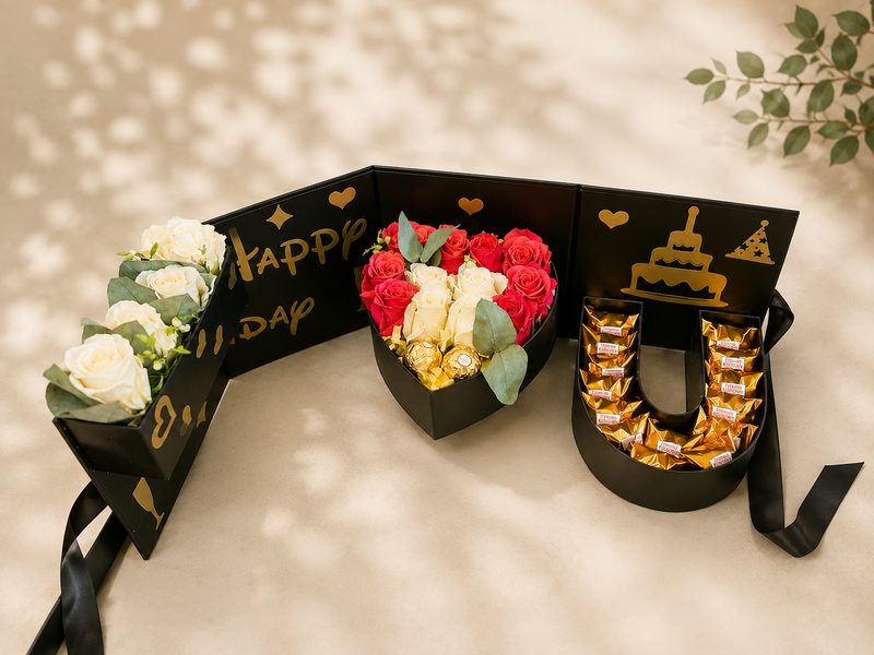 Drei schwarze Geschenkboxen mit goldenem Happy-Birthday-Schriftzug: längliche Box mit cremefarbenen Rosen, Herzbox mit roten und cremefarbenen Rosen und Ferrero Rocher, U-förmige Box mit Ferrero Küsschen